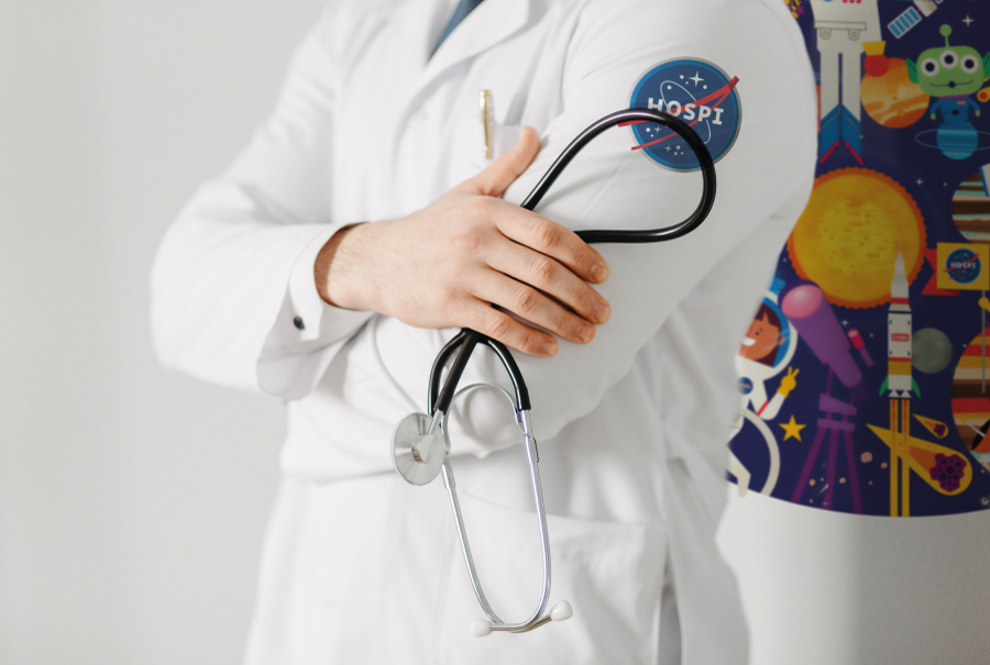Médico en bata blanca con un estetoscopio, destacando un parche de 'Estación Temporal Hospi' que adorna su bata, representando la temática espacial del hospital